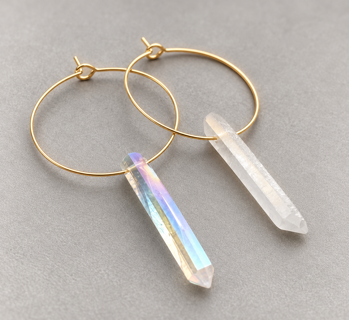 Gold hoop earrings with crystal point pendants, one in clear quartz and one in an iridescent rainbow crystal, displayed on a neutral gray background.