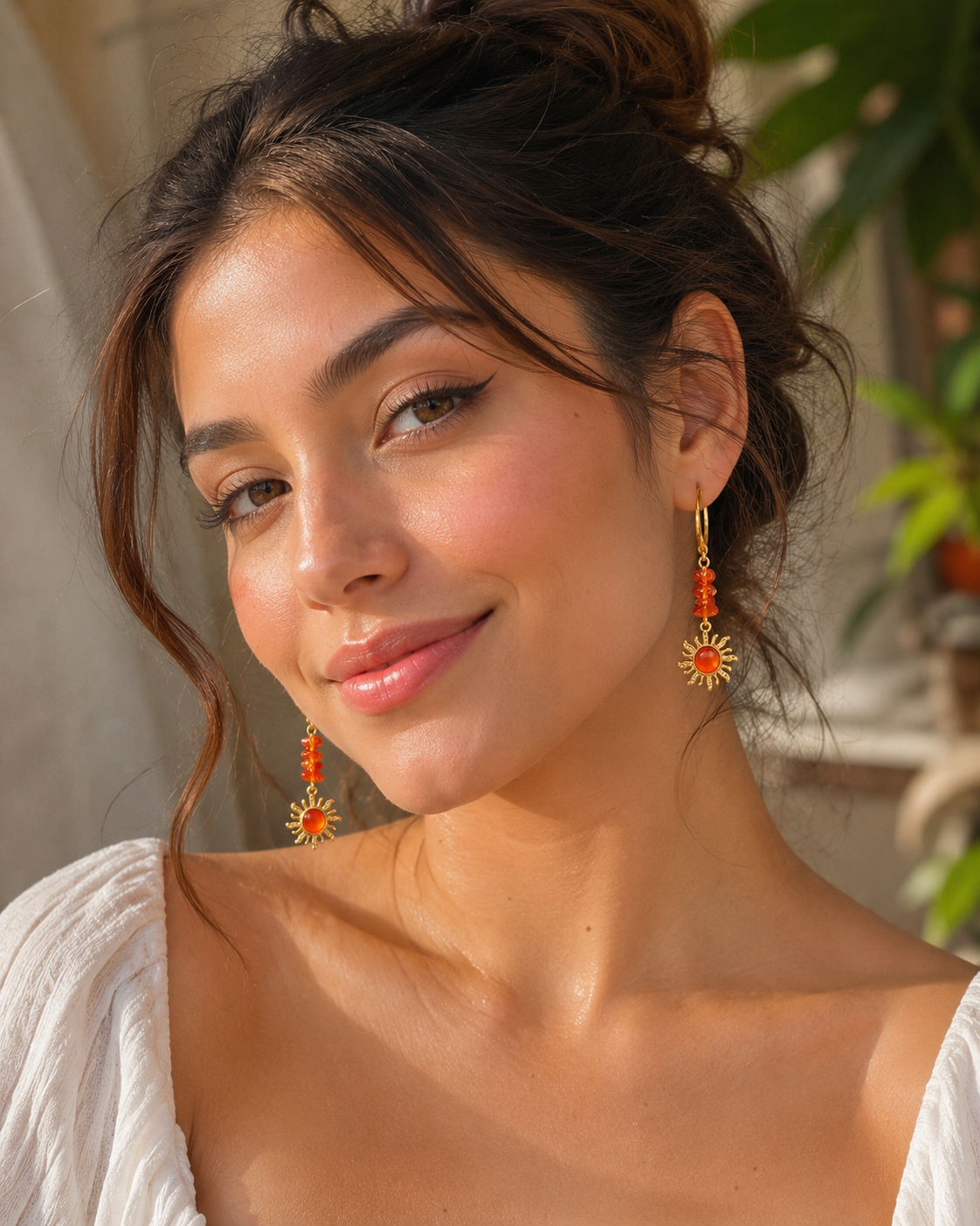 A woman with dark hair in a bun wearing dangling earrings with orange-red beads and gold sunburst charms, photographed in warm natural light against a softly blurred background.