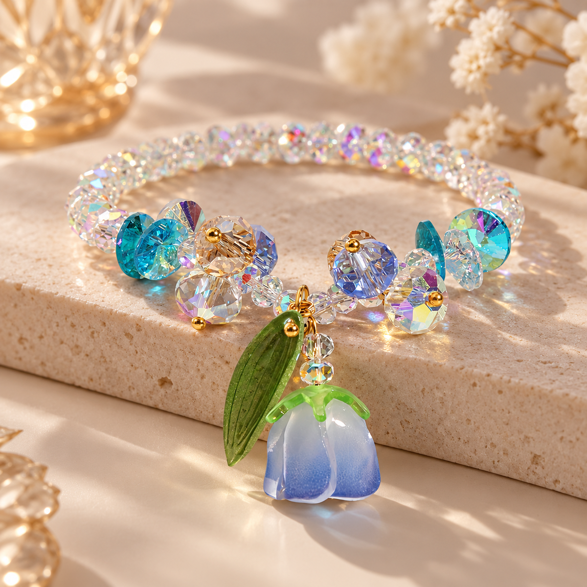 A beaded crystal bracelet with iridescent and blue faceted beads, gold-tone connectors, and a dangling flower charm in blue and green, displayed on a beige stone surface with soft decorative flowers in the background.