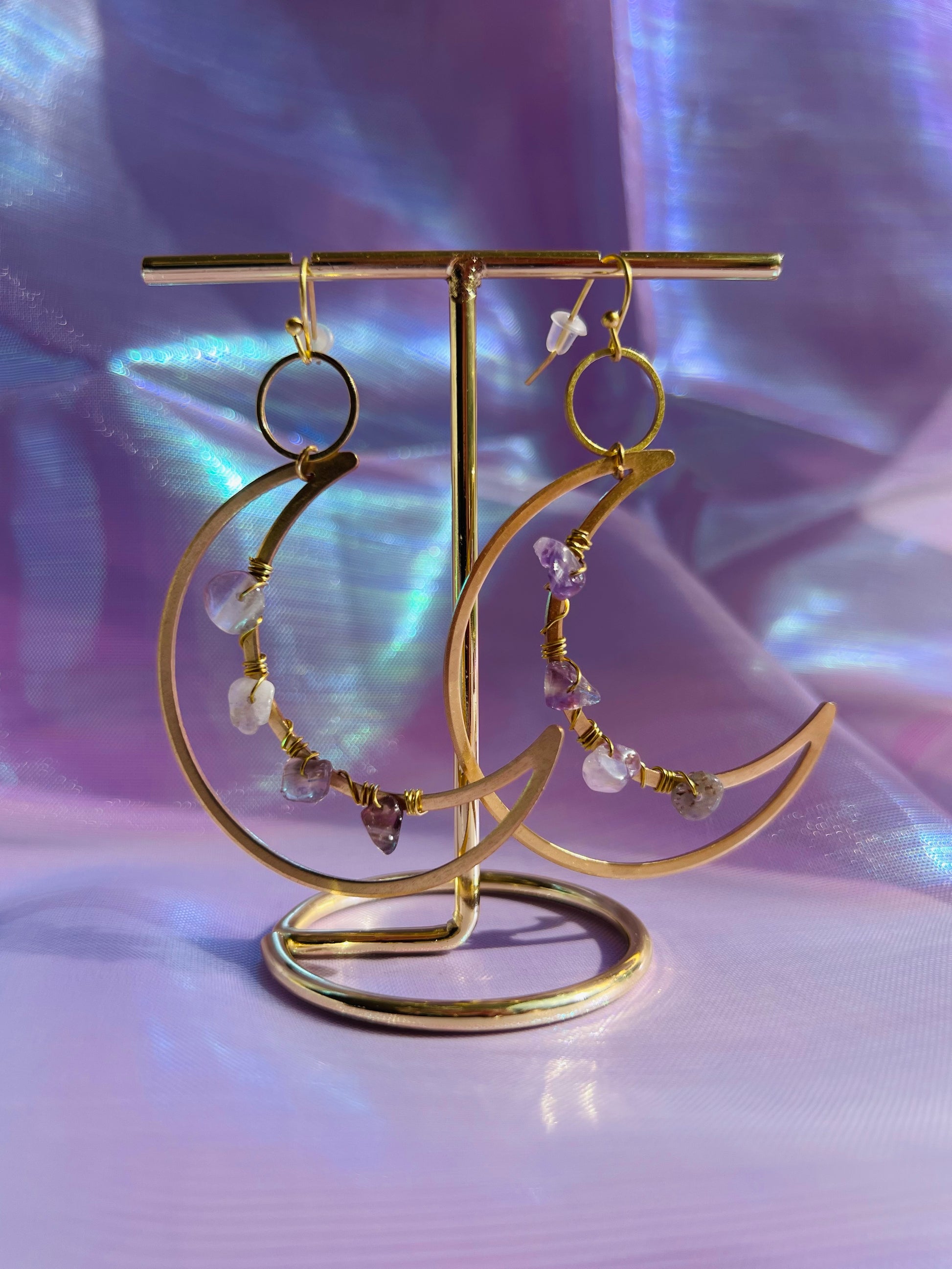A pair of gold-colored brass earrings in the shape of a crescent moon, featuring amethyst crystals and small dangling beads, displayed against a purple, glittery background.
