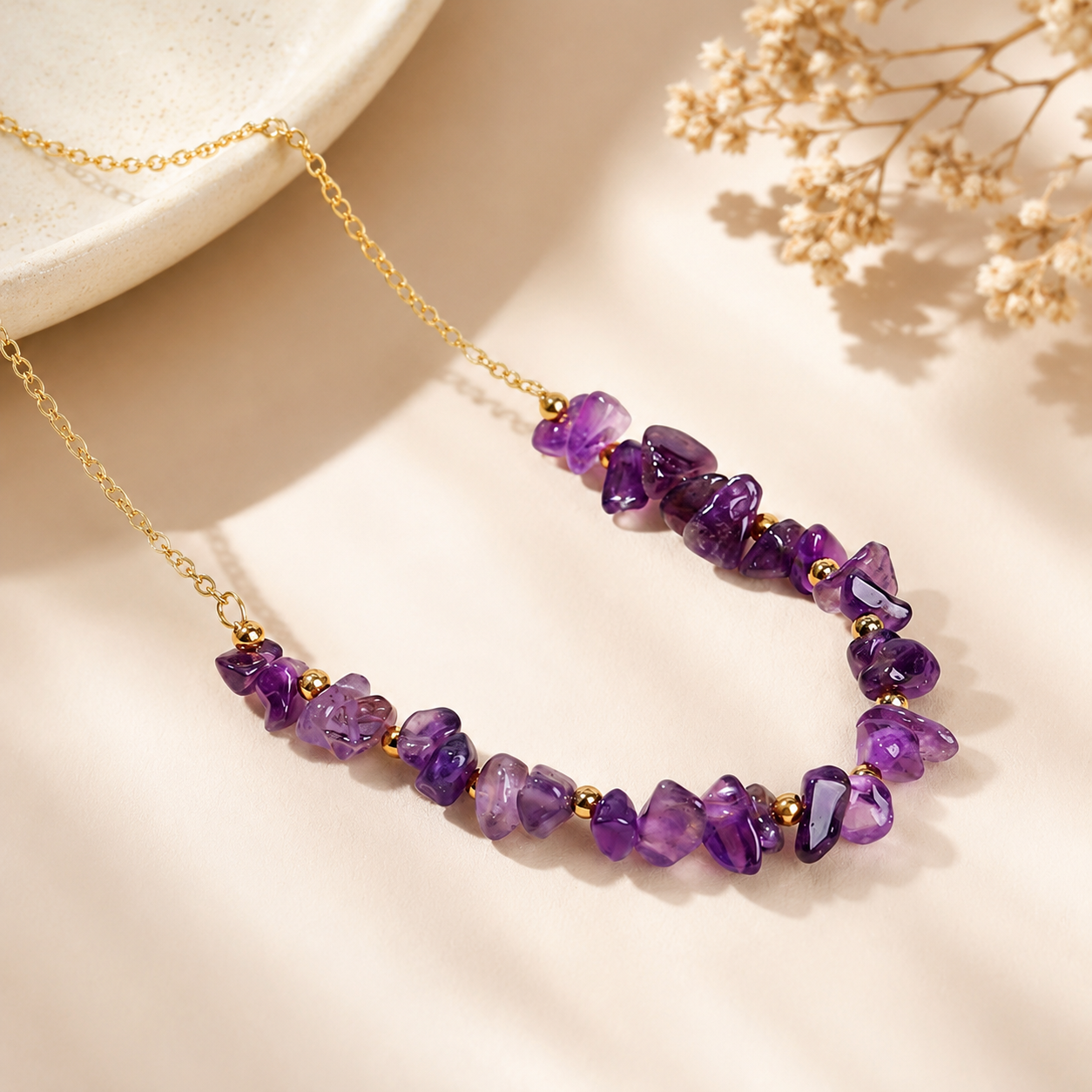 A gold chain necklace featuring irregular purple amethyst gemstone beads separated by small gold spacer beads, styled on a warm beige surface with dried flowers and a ceramic dish edge.