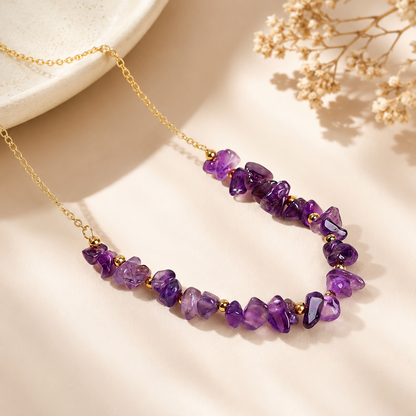A gold chain necklace featuring irregular purple amethyst gemstone beads separated by small gold spacer beads, styled on a warm beige surface with dried flowers and a ceramic dish edge.
