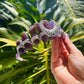 A hand holding a silver filigree hair claw clip decorated with purple amethyst crystals, set against a background of green tropical leaves.