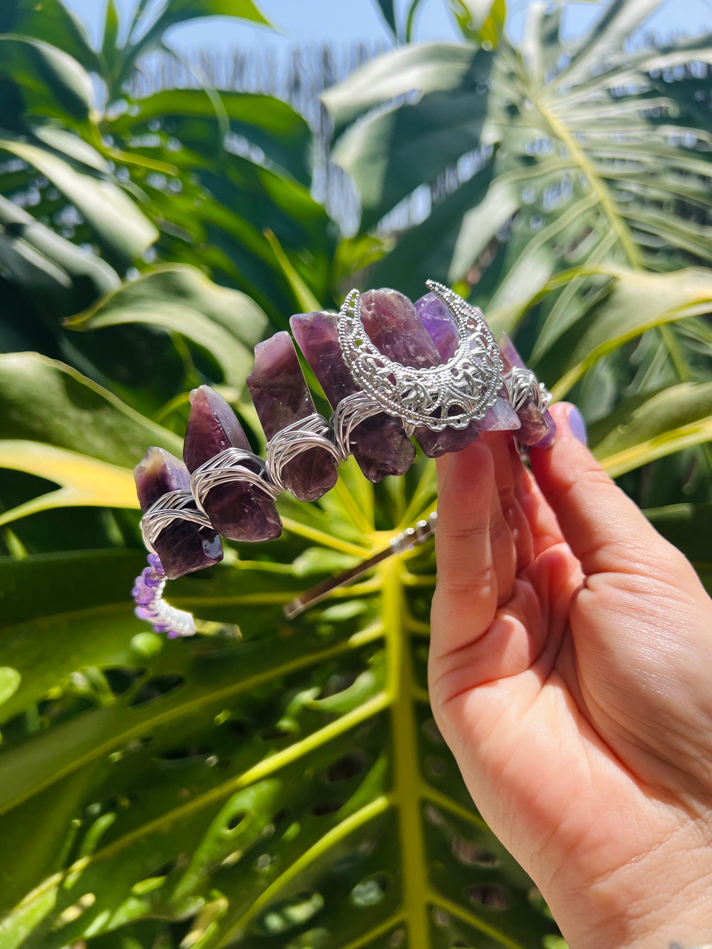 A hand holding a silver filigree hair claw clip decorated with purple amethyst crystals, set against a background of green tropical leaves.