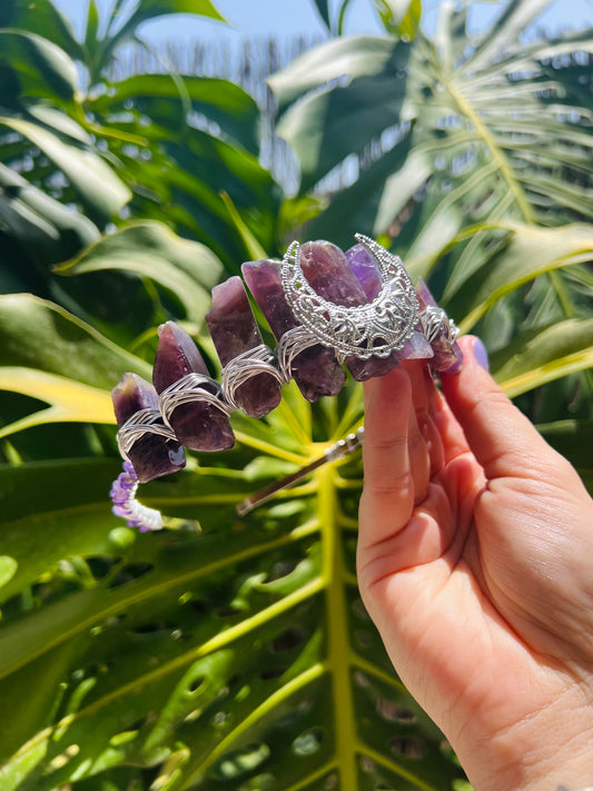 A hand holding a silver filigree hair claw clip decorated with purple amethyst crystals, set against a background of green tropical leaves.