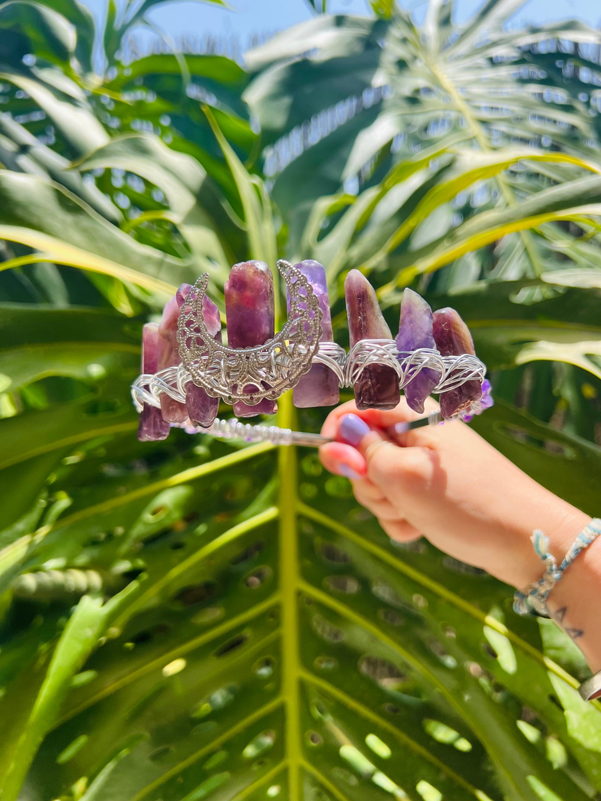 A hand holds a silver crescent moon tiara adorned with purple amethyst crystals against a background of large green monstera leaves.