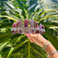 A silver filigree headband decorated with multiple purple amethyst crystal points, held up by a hand against a background of large green tropical monstera leaves.
