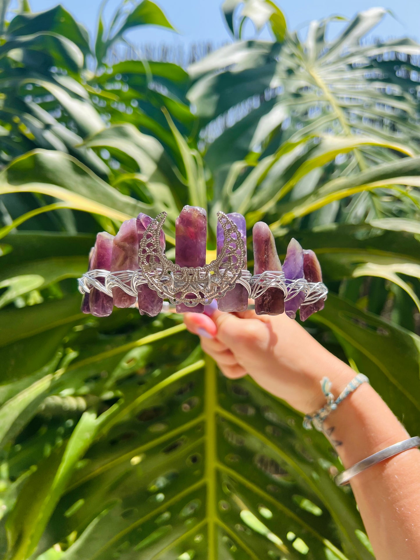 A silver filigree headband decorated with multiple purple amethyst crystal points, held up by a hand against a background of large green tropical monstera leaves.