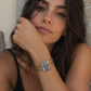 A close-up lifestyle shot of a woman wearing a stack of gold-toned bangles, including one with light blue stone accents, against a textured stone wall.