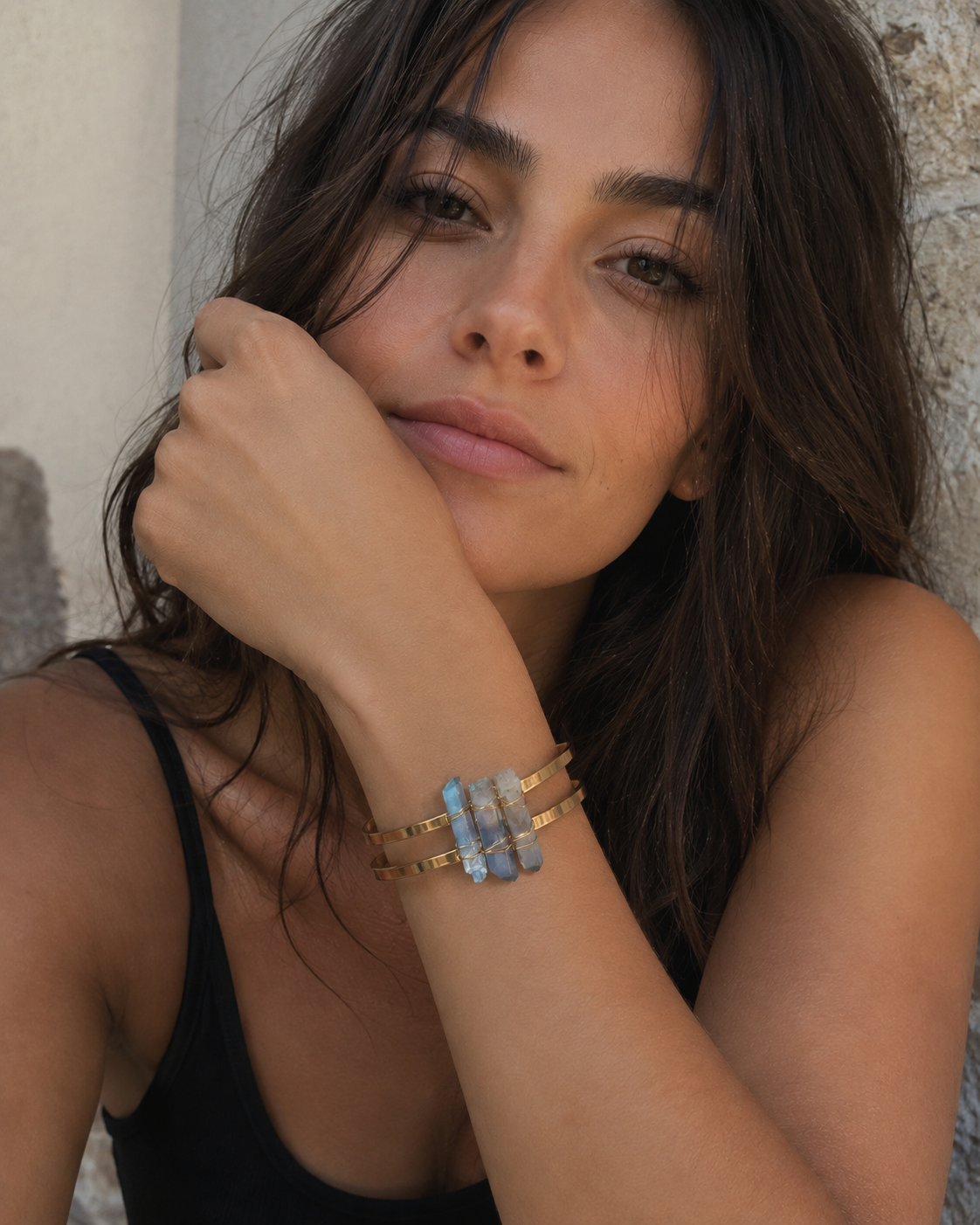 A close-up lifestyle shot of a woman wearing a stack of gold-toned bangles, including one with light blue stone accents, against a textured stone wall.