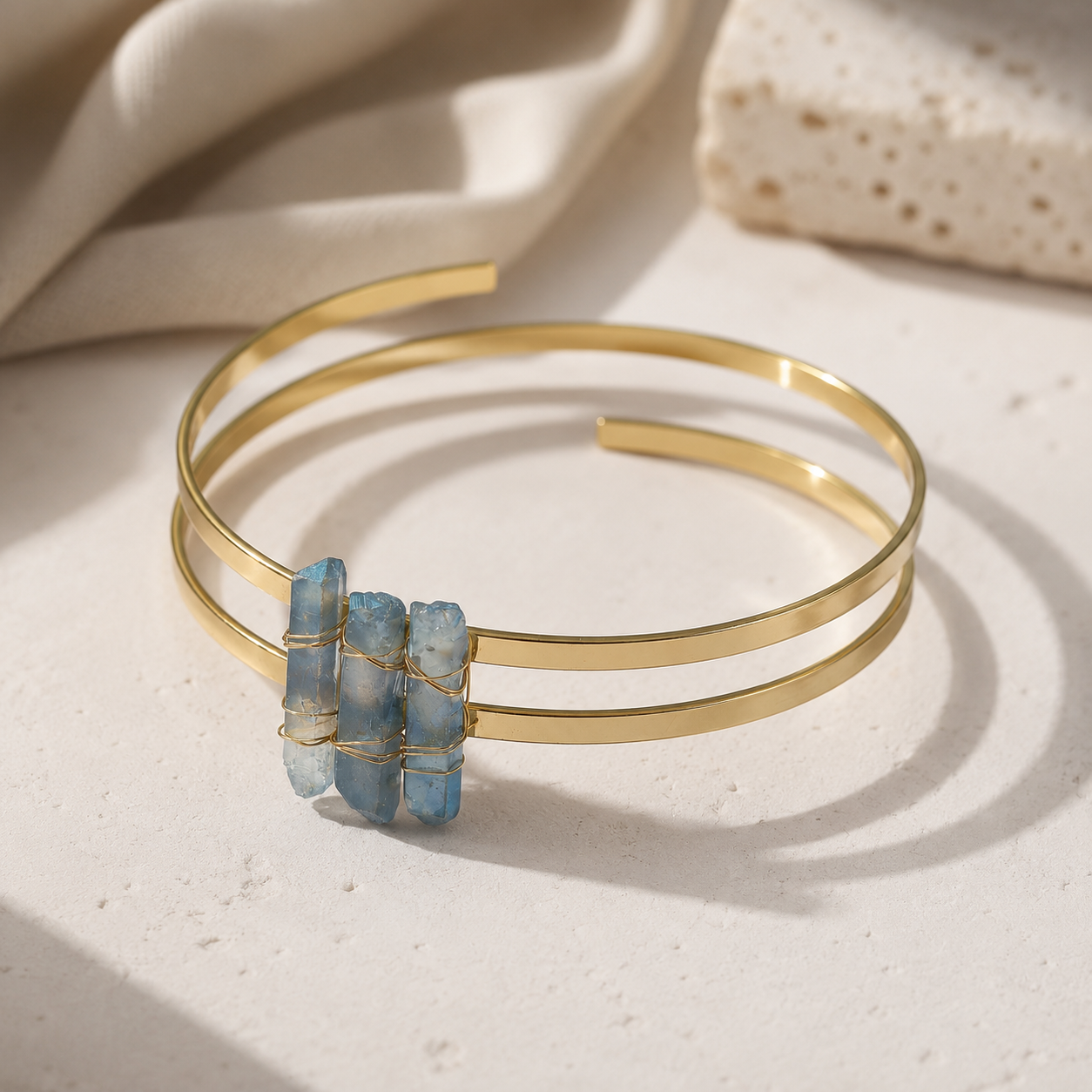 A double-band gold cuff bracelet featuring three small, stacked light blue stone charms wrapped in thin gold wire, displayed on a textured beige surface with soft fabric and stone props.