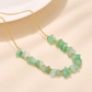 A gold chain necklace featuring a strand of polished light green stone chips separated by small gold beads, displayed on a soft beige surface.
