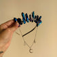 A hand holds up a celestial-themed headband featuring dark blue iridescent crystal points, a silver crescent moon, and a matching silver chain necklace with star and moon charms, set against a plain beige background.