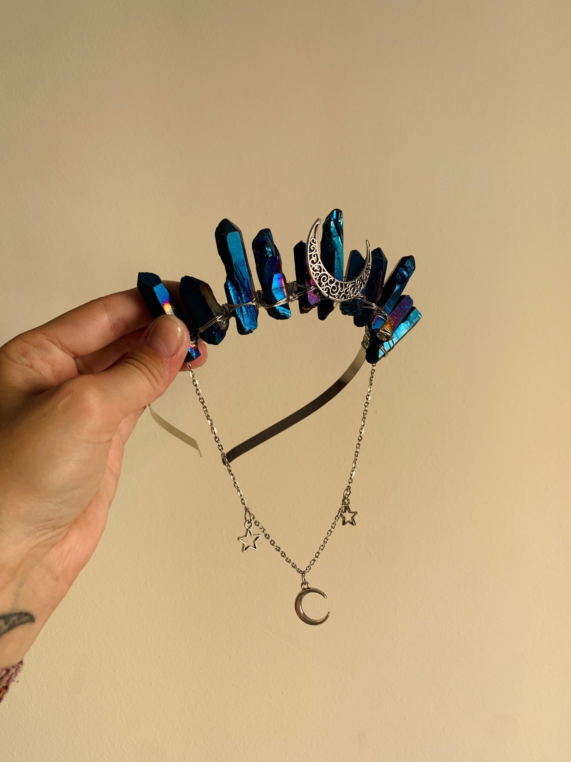 A hand holds up a celestial-themed headband featuring dark blue iridescent crystal points, a silver crescent moon, and a matching silver chain necklace with star and moon charms, set against a plain beige background.