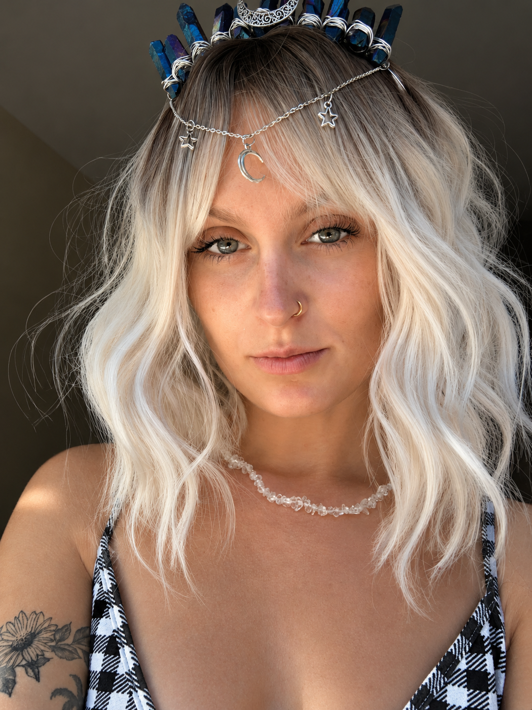 A close-up portrait of a woman with platinum blonde wavy hair wearing a silver crescent moon and star headpiece with blue and white crystal-like accents, along with a beaded necklace and a black-and-white patterned top.