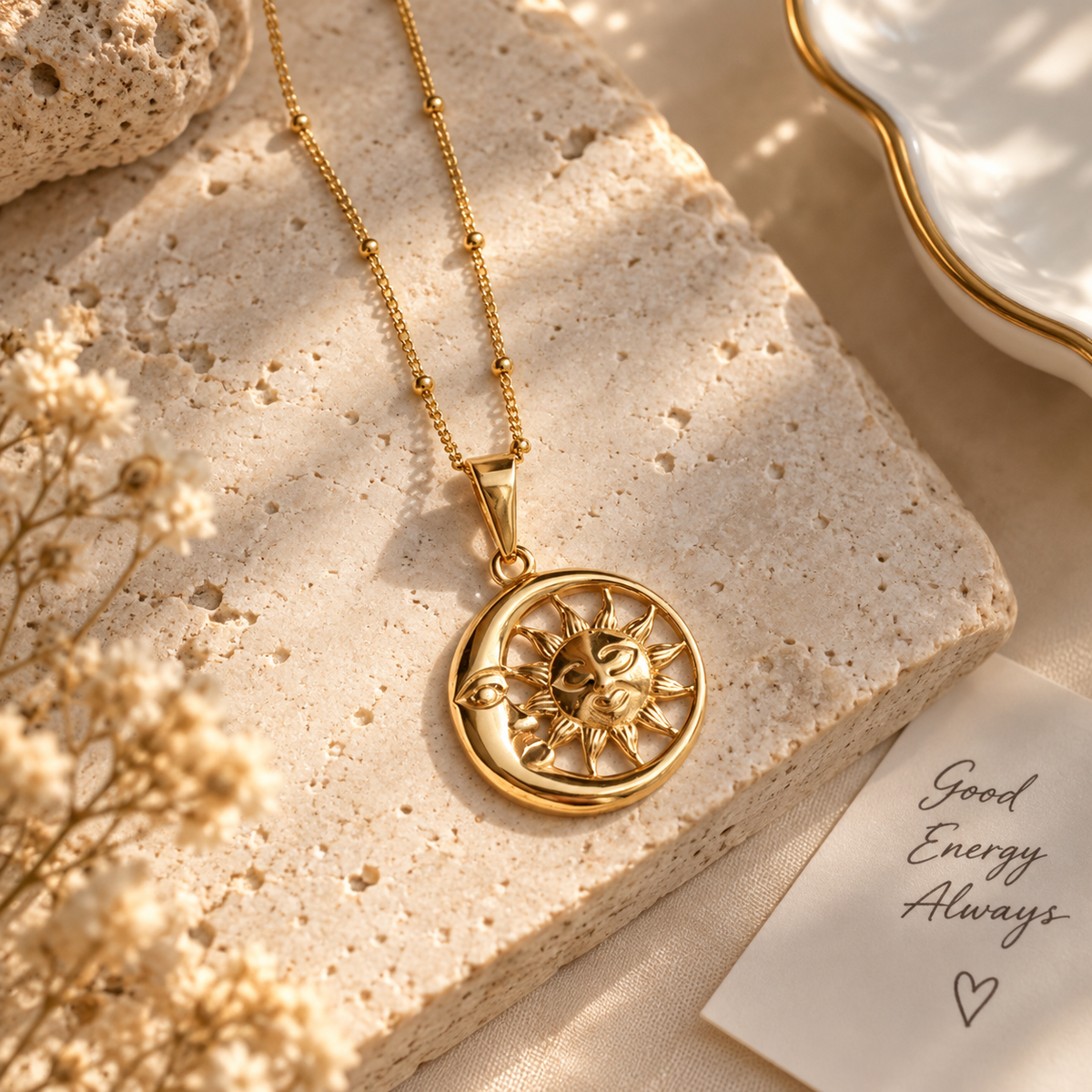 A gold pendant necklace featuring a circular sun and moon design, displayed on a textured beige stone block with dried flowers and a small card that reads 'Good Energy Always'.