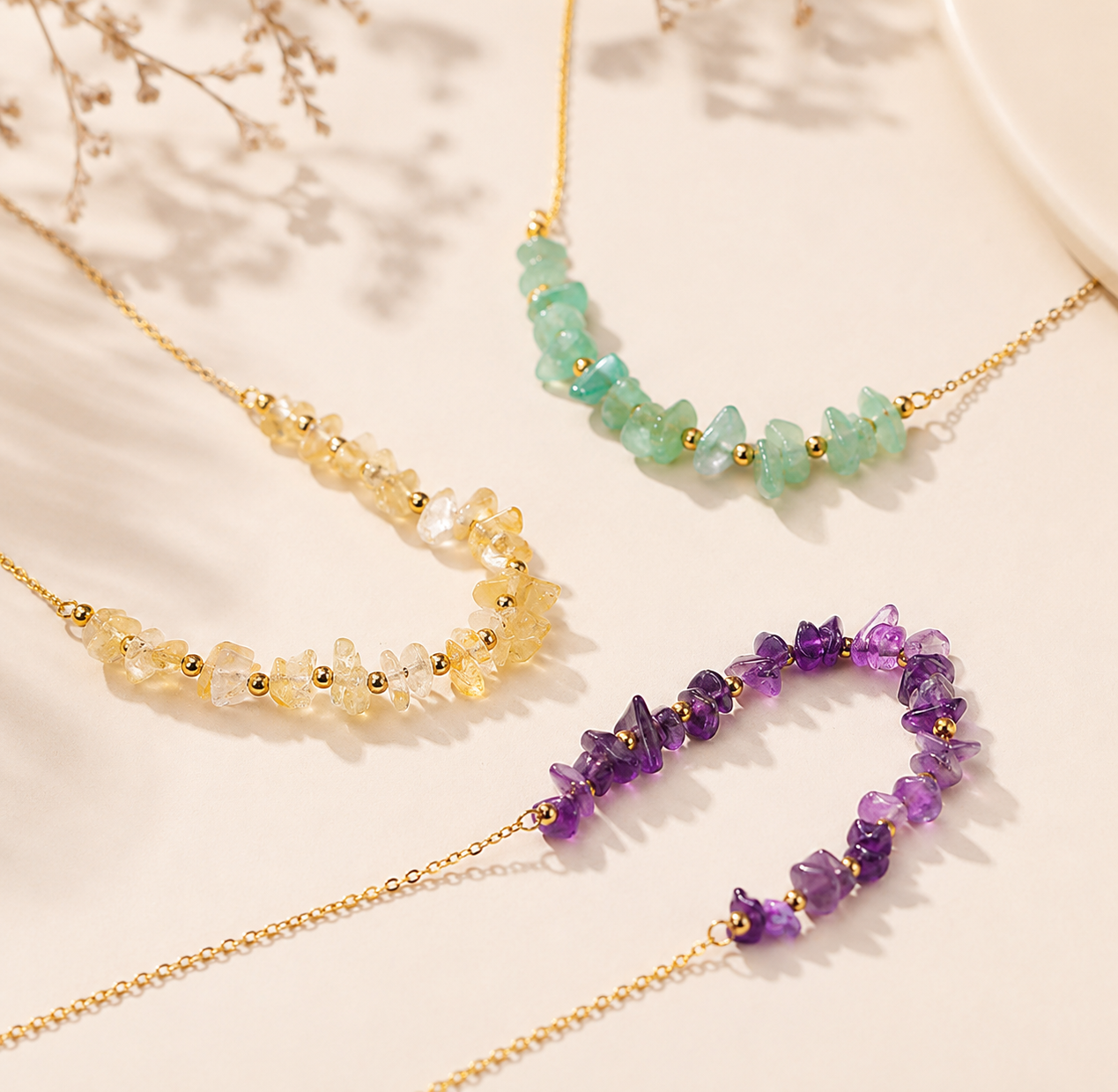 Three delicate gold chain necklaces with chunky gemstone beads in yellow, light green, and purple, arranged on a light beige background with soft shadows.