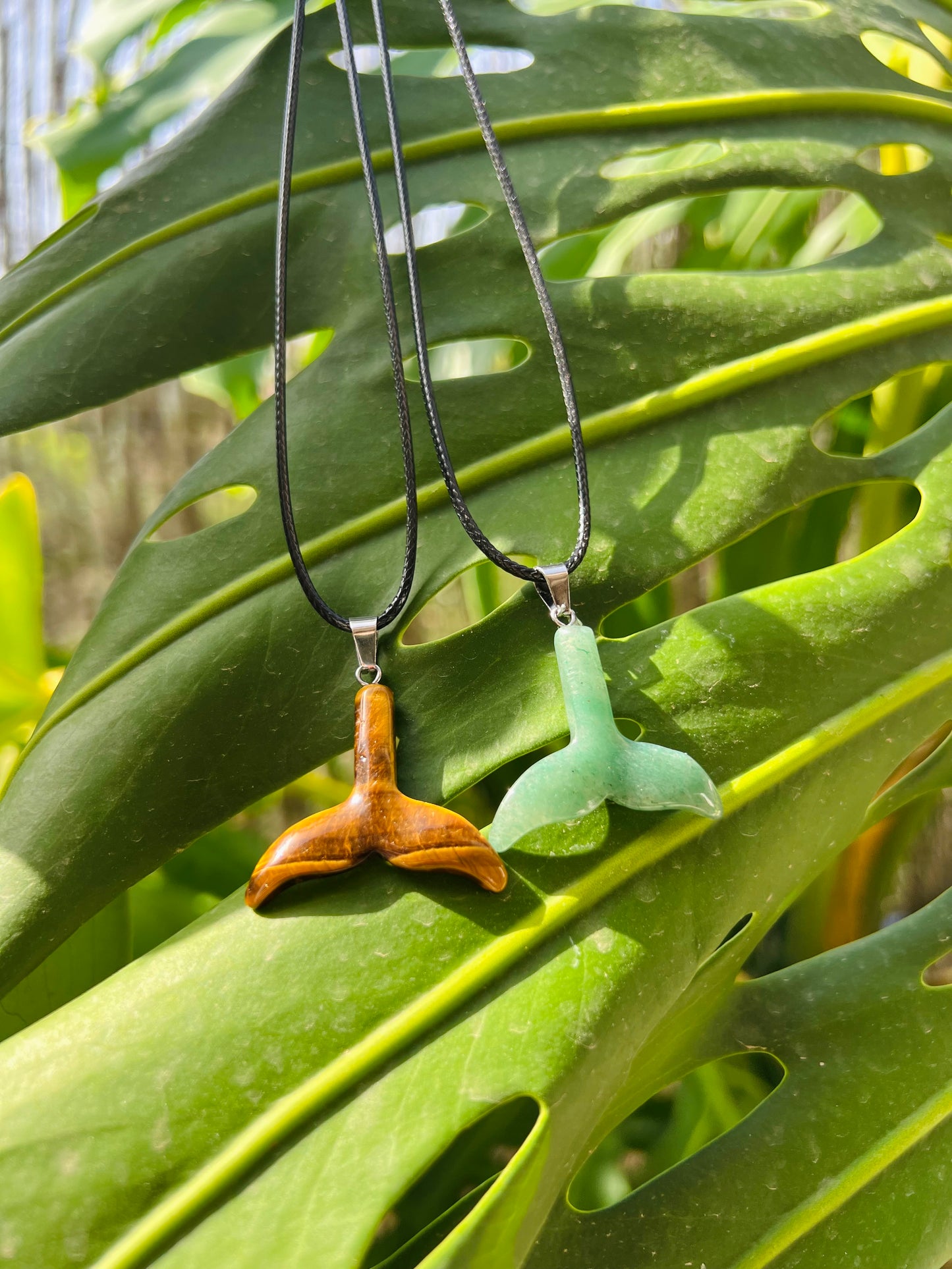 Two whale tail pendant necklaces hanging against a large green monstera leaf. One pendant is carved from brown wood and the other from light green stone, both on black cords.