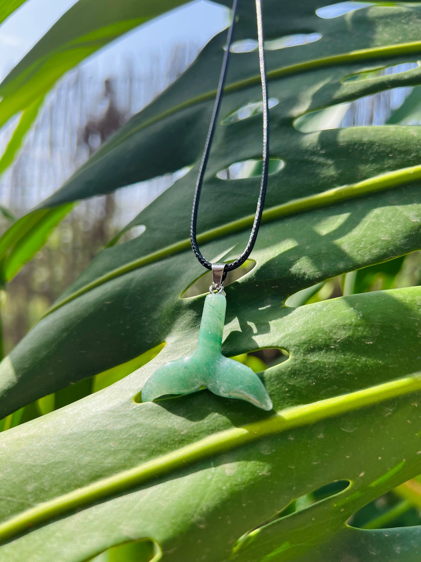 A green jade pendant shaped like a mermaid tail hanging from a black cord necklace, set against a background of large green monstera leaves.