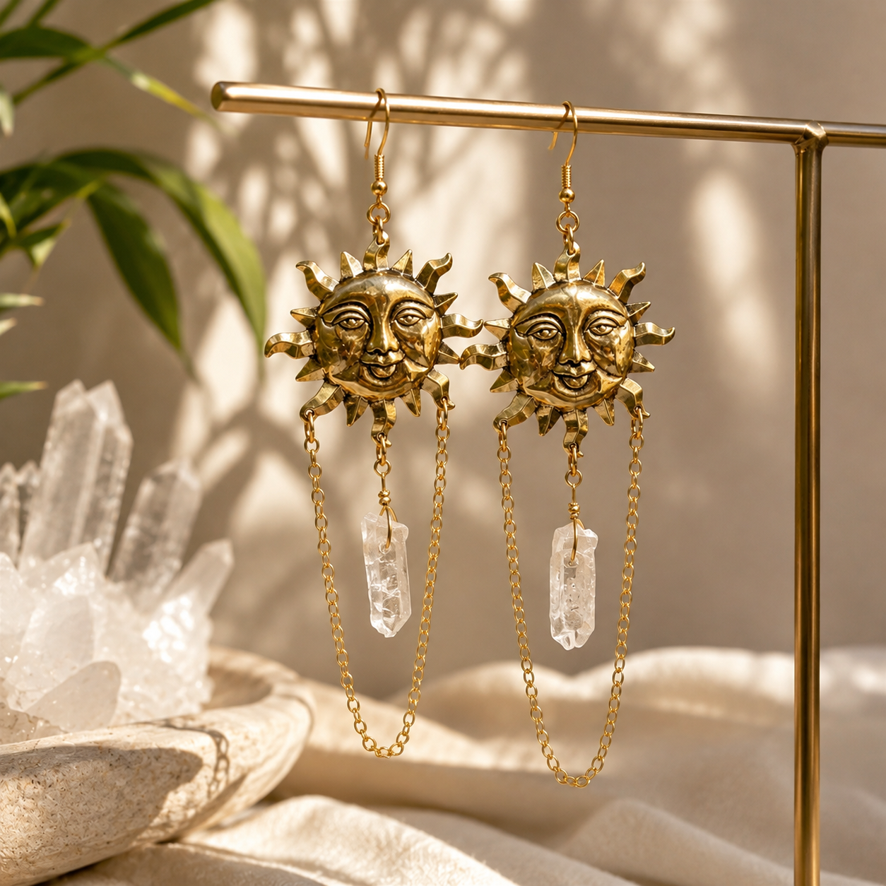 A pair of gold sun-face dangle earrings with clear crystal drops, hanging from a gold display stand against a warm beige background with soft shadows and decorative crystals.