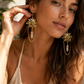 A close-up lifestyle shot of a woman wearing ornate gold-toned dangle earrings with sunburst details and clear crystal-like drops, paired with a cream crochet top.