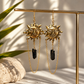 A pair of gold-toned dangling earrings with sun-face charms and black rectangular stone pendants, hanging from a gold display stand in a styled setting with soft shadows and a plant leaf in the background.