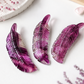 Three carved feather-shaped jewelry pieces in translucent purple and magenta tones arranged on a white surface with soft decorative props.