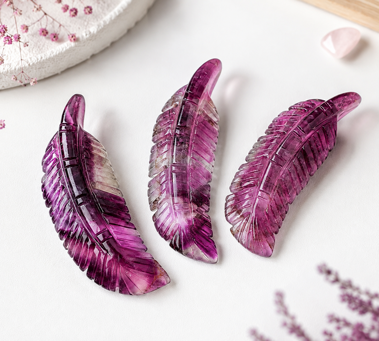Three carved feather-shaped jewelry pieces in translucent purple and magenta tones arranged on a white surface with soft decorative props.