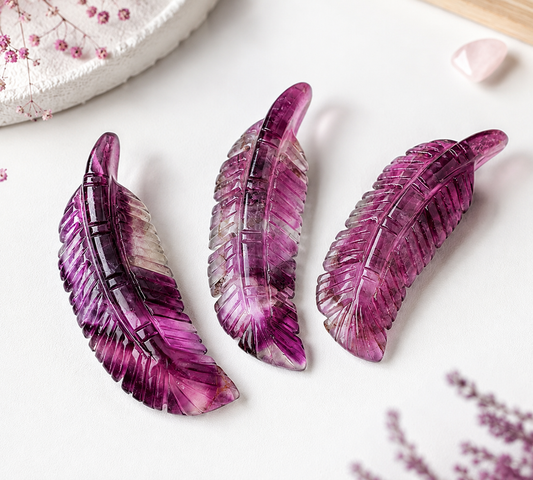 Three carved feather-shaped jewelry pieces in translucent purple and magenta tones arranged on a white surface with soft decorative props.