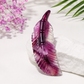 A carved purple amethyst feather-shaped pendant displayed on a white textured stone, with pink flowers and green palm leaves in the background.