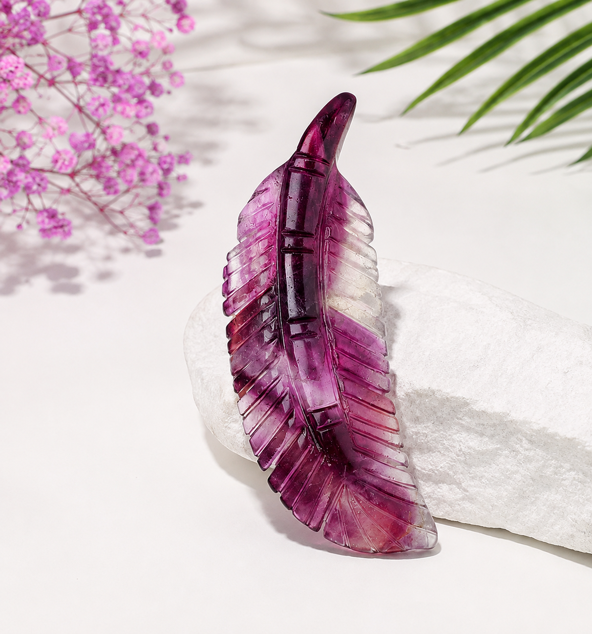 A carved purple amethyst feather-shaped pendant displayed on a white textured stone, with pink flowers and green palm leaves in the background.