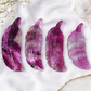 Four carved feather-shaped crystal pendants in shades of purple and magenta, arranged on a white fabric background with dried flowers and a crystal cluster.