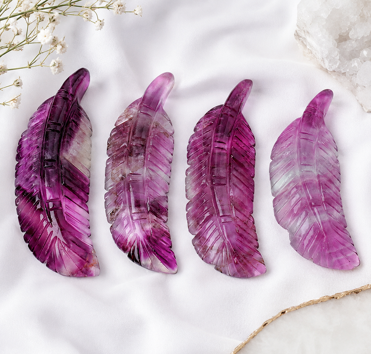 Four carved feather-shaped crystal pendants in shades of purple and magenta, arranged on a white fabric background with dried flowers and a crystal cluster.