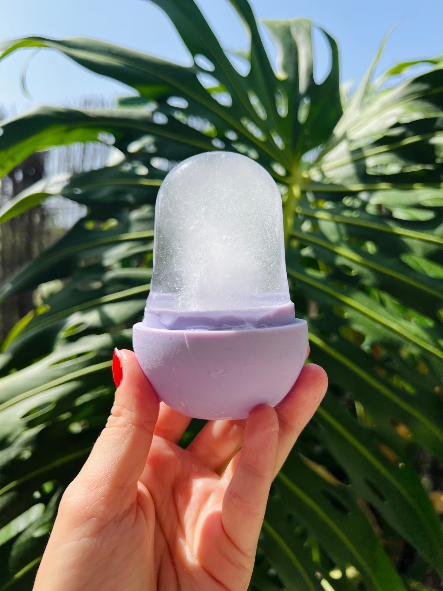A hand with red nail polish holds a lavender purple silicone mold containing a clear ice cube, set against a background of large green monstera leaves and blue sky.