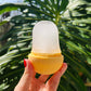 A hand with red nail polish holds a yellow and frosted ice mold against a background of large green monstera leaves and blue sky.