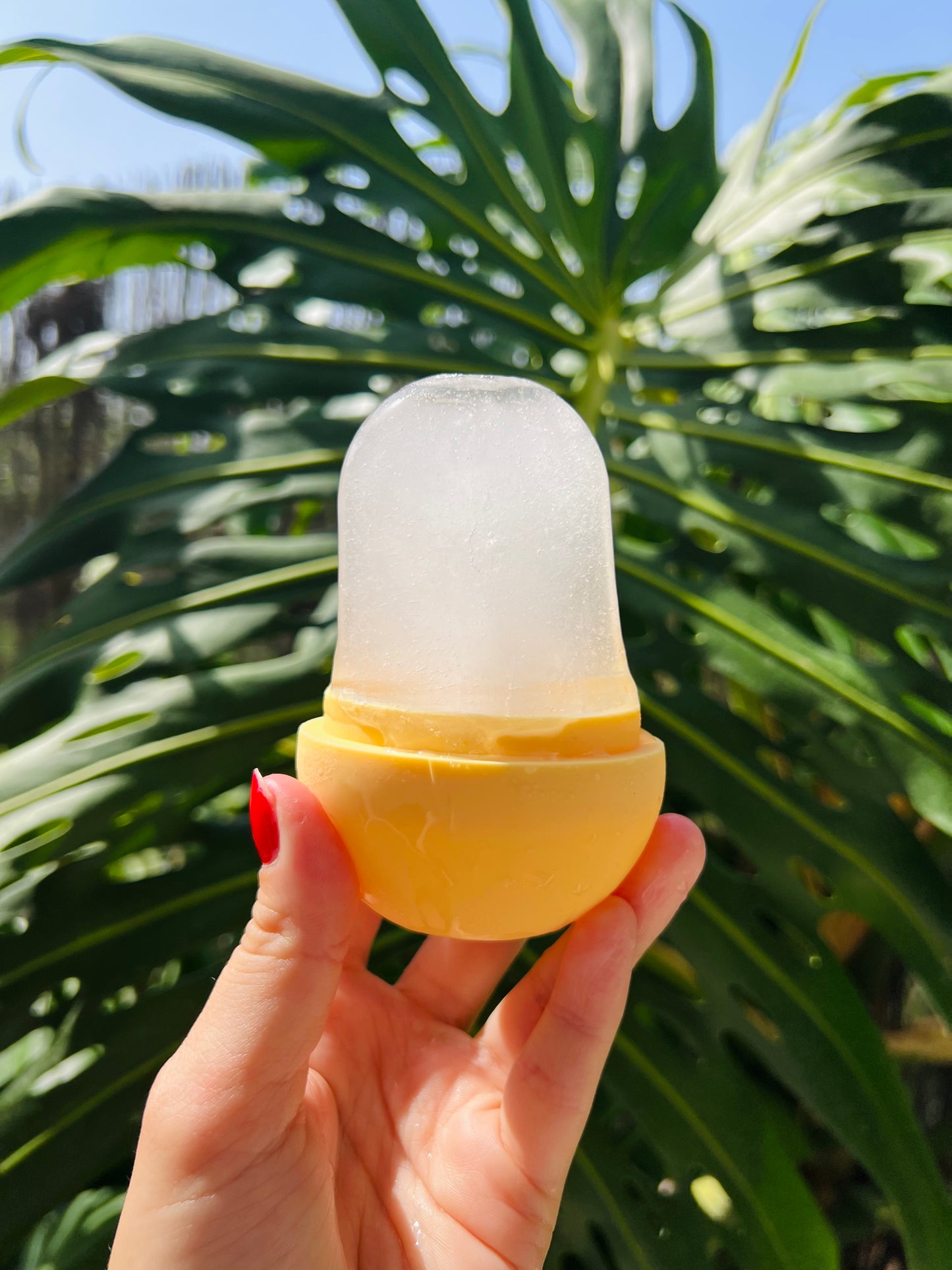 A hand with red nail polish holds a yellow and frosted ice mold against a background of large green monstera leaves and blue sky.