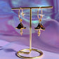 A pair of gold-colored mushroom-shaped earrings displayed on a stand against a purple and blue background.