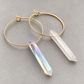 Gold hoop earrings with crystal point pendants, one in clear quartz and one in an iridescent rainbow crystal, displayed on a neutral gray background.