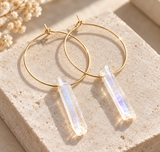 Gold hoop earrings with clear iridescent crystal point pendants, displayed on a textured beige stone block with dried flowers in the background.
