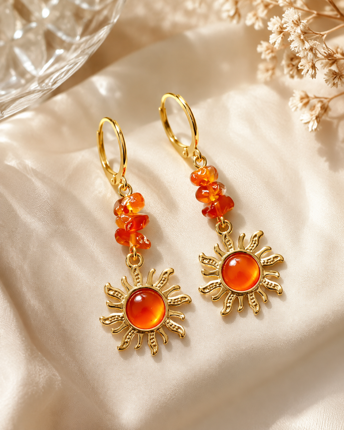A pair of gold drop earrings with clustered orange beads and sun-shaped charms featuring round orange cabochon stones, displayed on draped cream satin fabric with dried flowers and a glass bowl in the background.