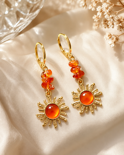 A pair of gold drop earrings with clustered orange beads and sun-shaped charms featuring round orange cabochon stones, displayed on draped cream satin fabric with dried flowers and a glass bowl in the background.