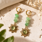 A pair of bohemian-style dangle earrings with gold-toned hoops, green faceted stone beads, and sunburst charms with oval green centers, displayed on a textured beige stone surface with dried flowers and green leaves.