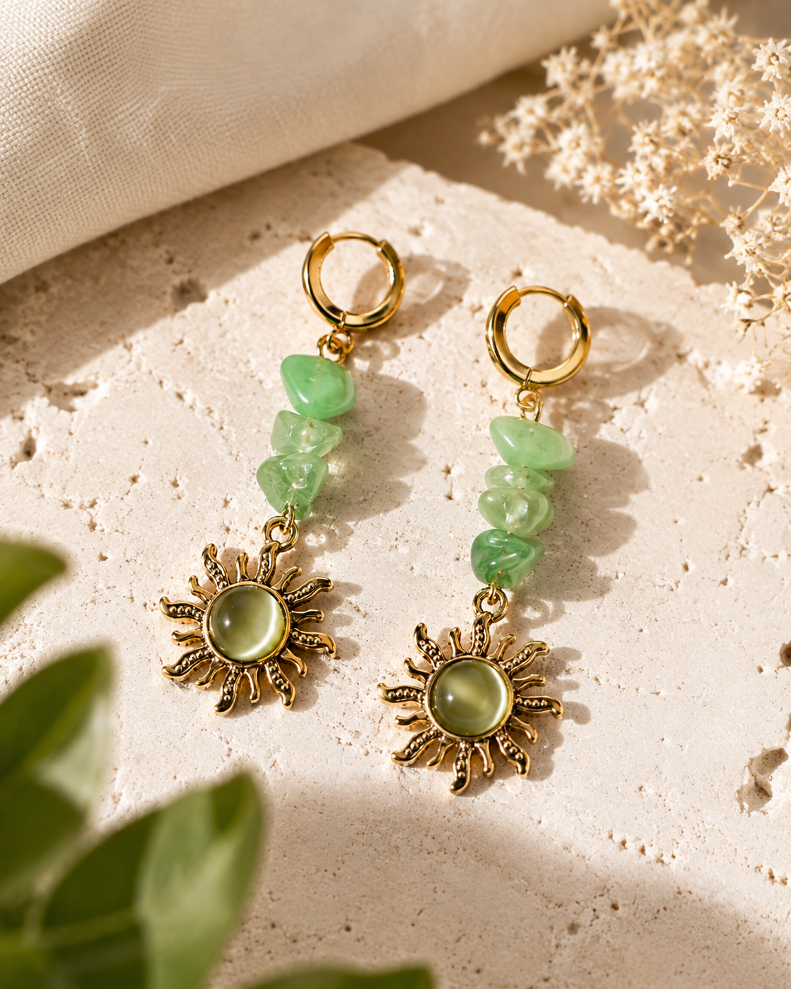 A pair of bohemian-style dangle earrings with gold-toned hoops, green faceted stone beads, and sunburst charms with oval green centers, displayed on a textured beige stone surface with dried flowers and green leaves.