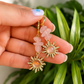 A pair of gold-tone dangle earrings with pink rose quartz stone accents and sunburst charms, held in a hand with green foliage in the background.