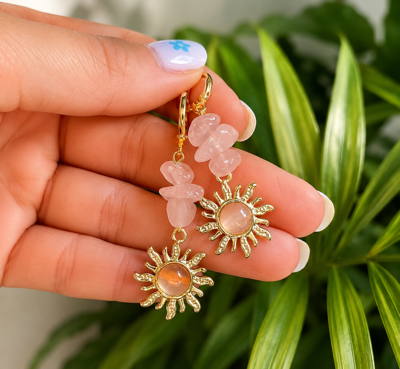 A pair of gold-tone dangle earrings with pink rose quartz stone accents and sunburst charms, held in a hand with green foliage in the background.