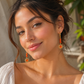 A woman with dark hair in a bun wearing dangling earrings with orange-red beads and gold sunburst charms, photographed in warm natural light against a softly blurred background.