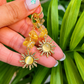 A pair of gold-toned dangle earrings with yellow crystal beads and sun-shaped charms, held in a hand against a background of green plant leaves.