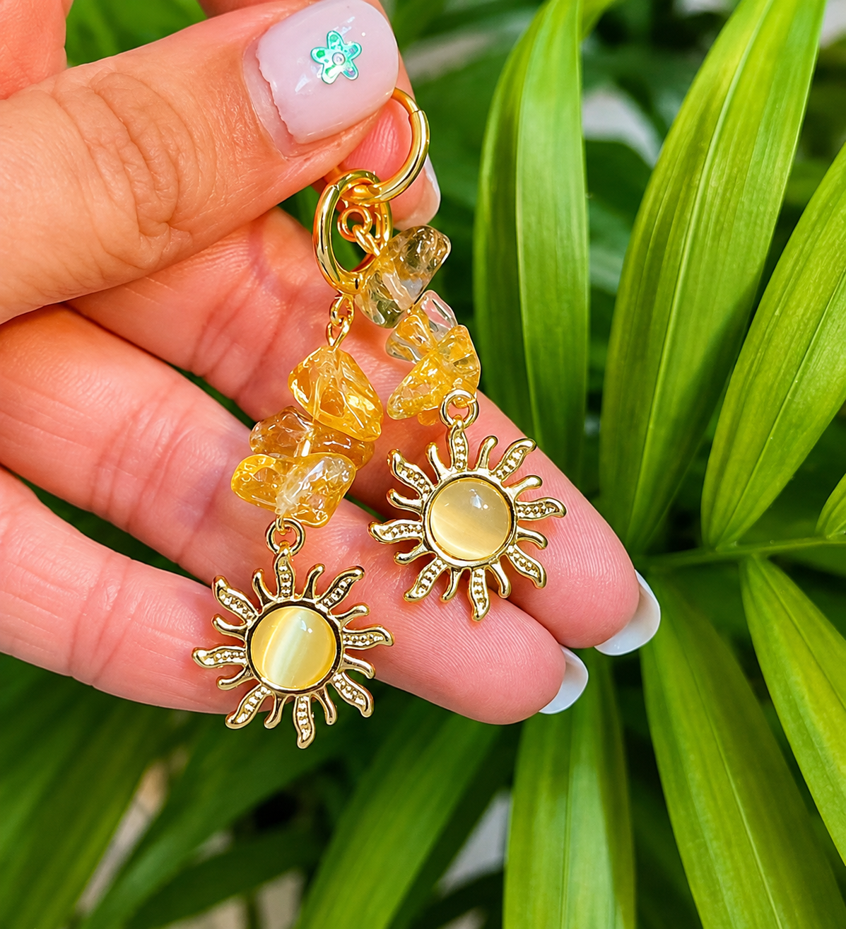 A pair of gold-toned dangle earrings with yellow crystal beads and sun-shaped charms, held in a hand against a background of green plant leaves.