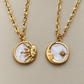 Two gold-tone pendant necklaces on matching chains, displayed side by side on a neutral beige background. The left necklace features a circular pendant with a gold sun face set against an iridescent white mother-of-pearl background.