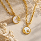 Two gold-tone chain necklaces with oval mother-of-pearl pendants, one featuring a sun motif and the other a crescent moon with a star, styled on a beige stone and dried flowers.